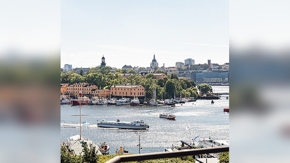 Stiligt - Utsikt över en stadsbild vid vattnet med båtar på vattnet, historiska byggnader, träd och avlägsna moderna byggnader under en klar himmel.