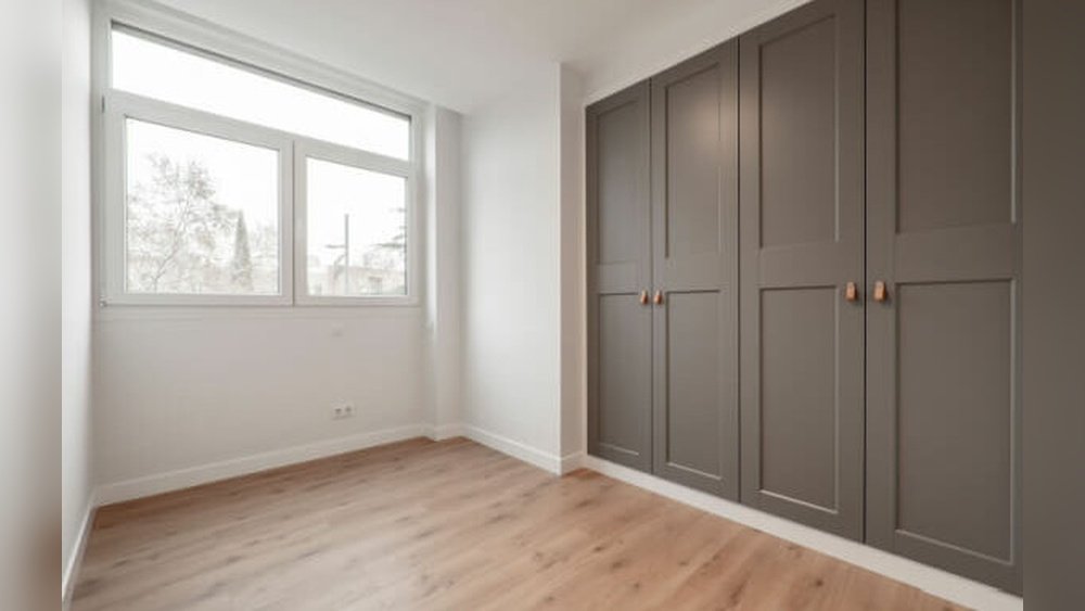 Stiligt - Tomt rum med ett stort fönster, ljust trägolv, vita väggar och inbyggda grå garderober med kopparhandtag.