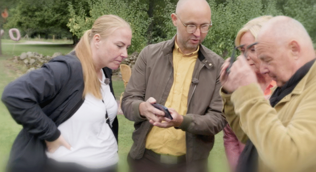 Stiligt - Fyra vuxna står utomhus och tittar noga på en smartphone som en av dem håller i. I bakgrunden syns träd och grönska.