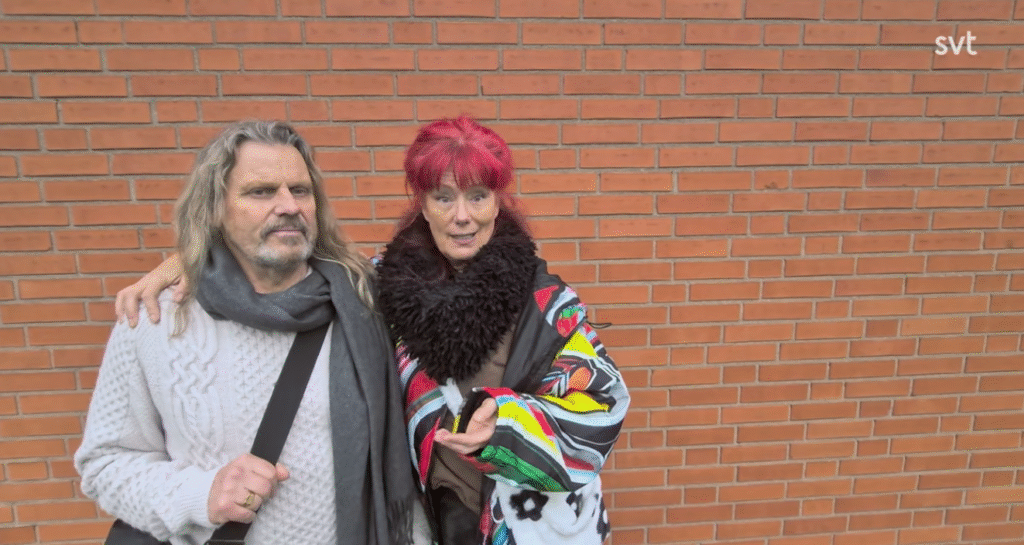 Stiligt - Två vuxna står sida vid sida framför en tegelvägg; den ena har långt hår och skägg, den andra har ljusrött hår och bär en färgglad sjal.