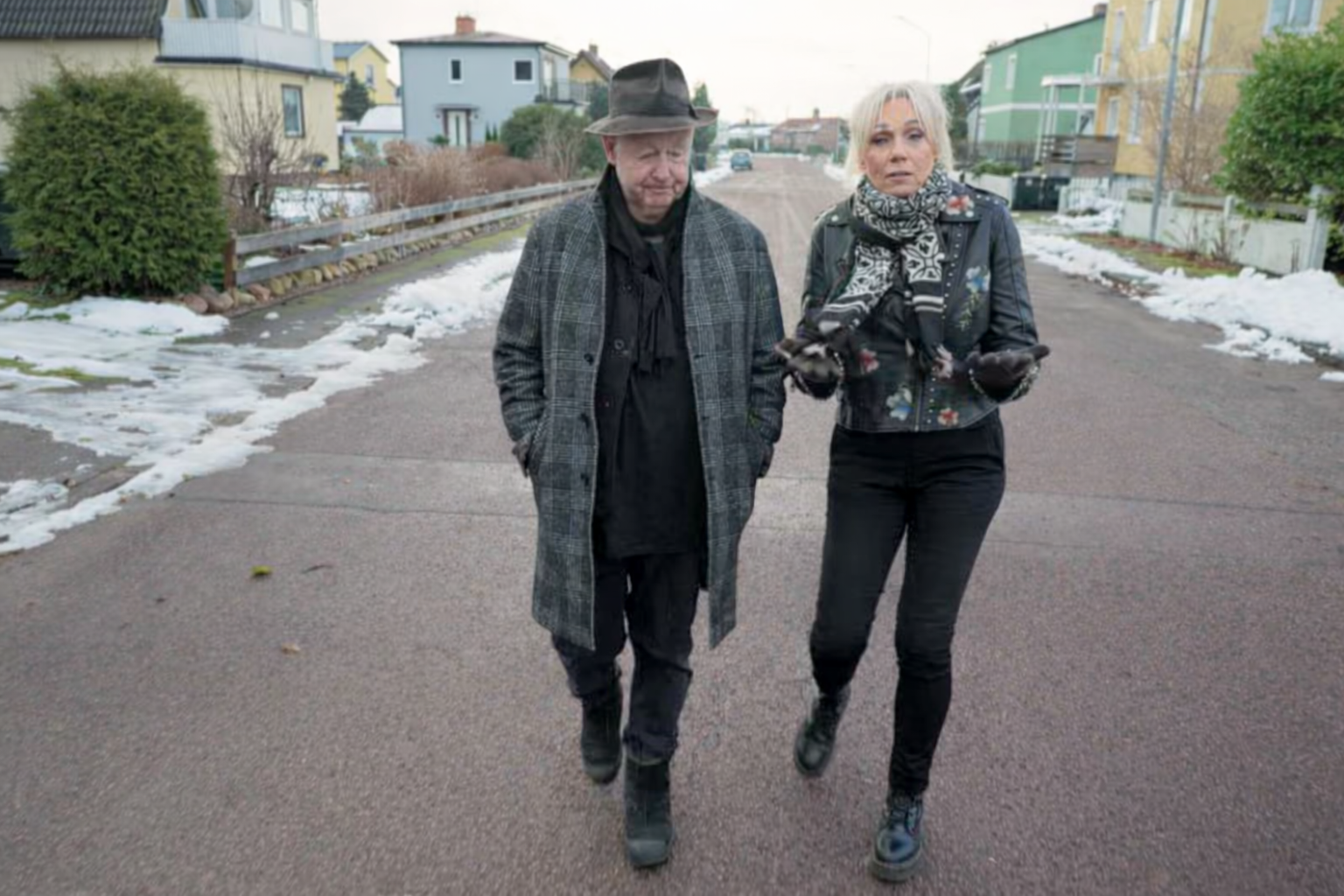Stiligt - Två personer, den ena klädd i rutig rock och hatt och den andra i svart jacka och halsduk, går tillsammans på en snöig villagata.