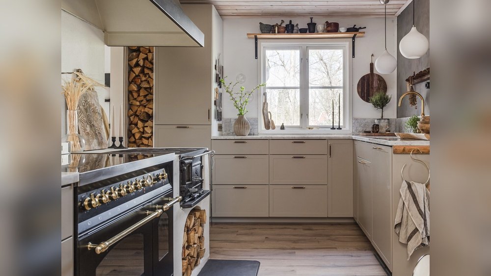 Stiligt - Modernt kök med beige skåp, en spis i svart och guld, staplad ved, trädetaljer och ett stort fönster som släpper in naturligt ljus.