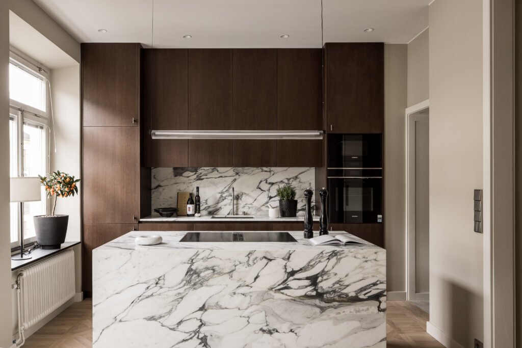 Stiligt - Modernt kök med skåp i mörkt trä, köksö och stänkskydd i marmor, inbyggda vitvaror och krukväxter nära ett fönster.