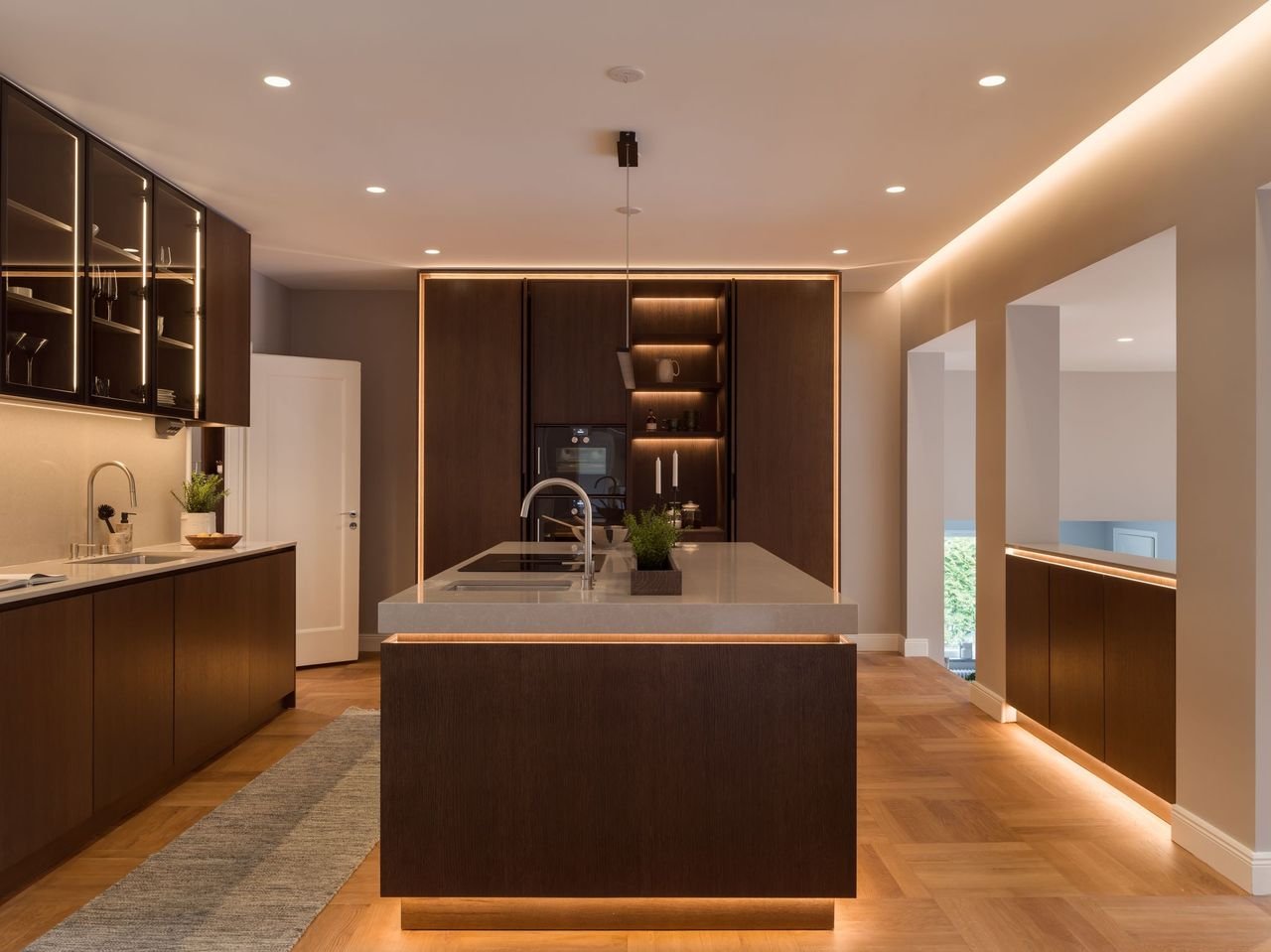 Stiligt - Modernt kök med skåp i mörkt trä, en central ö med diskho, inbyggda vitvaror, belysning under skåpen och trägolv.