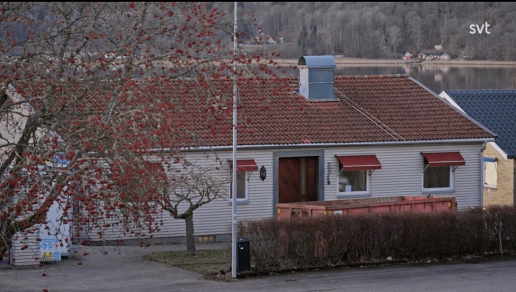 Stiligt - Ett enplanshus med rött tegeltak, röda markiser och en stor sopcontainer på framsidan, beläget vid en sjö med träd i bakgrunden.