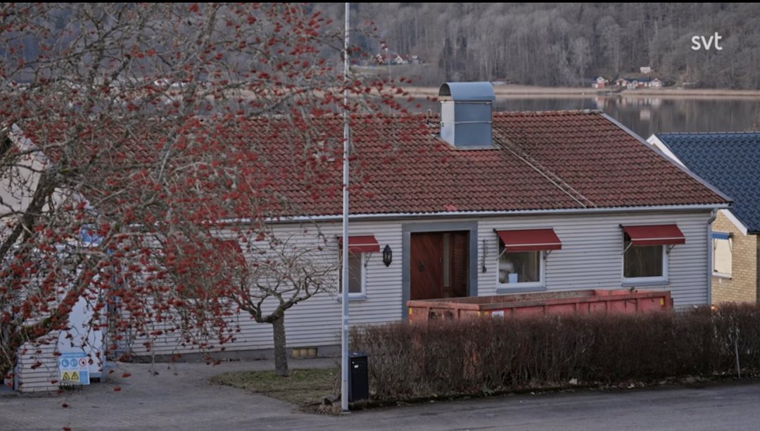 Stiligt - Ett enplanshus med rött tegeltak, röda markiser och en stor sopcontainer på framsidan, beläget vid en sjö med träd i bakgrunden.