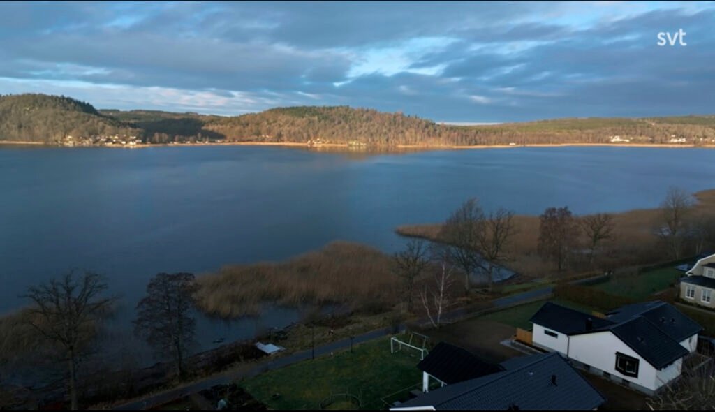 Stiligt - Ett sjölandskap med lugnt vatten, omgivande kullar och flera hus nära strandlinjen under en delvis molnig himmel.