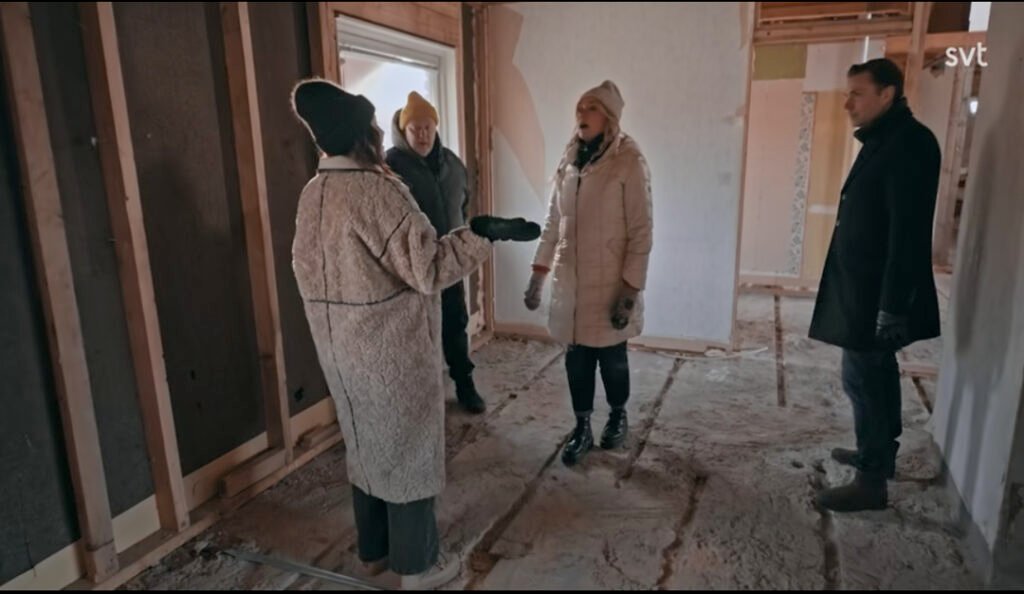 Stiligt - Fyra personer klädda i vinterkläder står och pratar i en ofärdig byggnad med synliga väggar och golv.