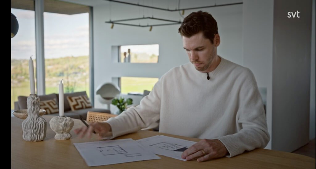 Stiligt - En man i vit tröja sitter vid ett bord och går igenom arkitektritningar i ett modernt, väl upplyst rum med stora fönster och dekorativa ljus.
