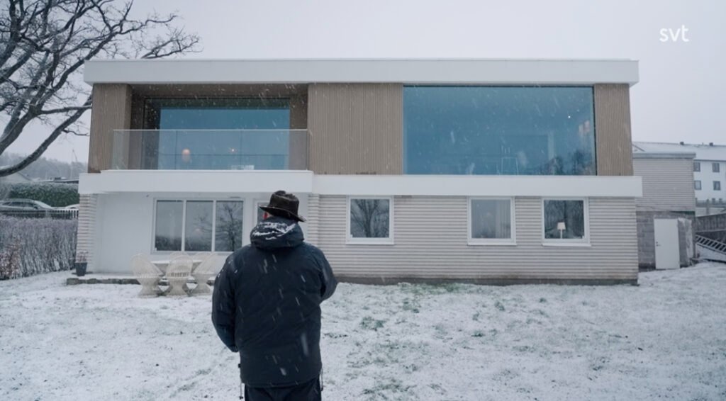 Stiligt - En person i vinterkläder står framför ett modernt tvåvåningshus med stora fönster under snöfall.