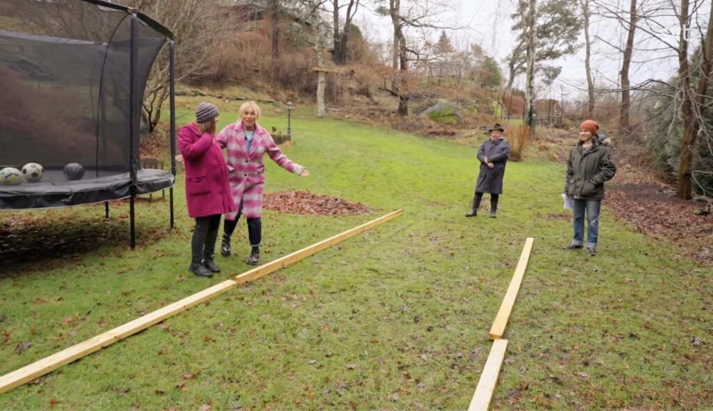 Stiligt - Fyra personer står i gräset nära två parallella träbjälkar, till vänster en studsmatta med bollar och i bakgrunden träd och hus under en mulen dag.