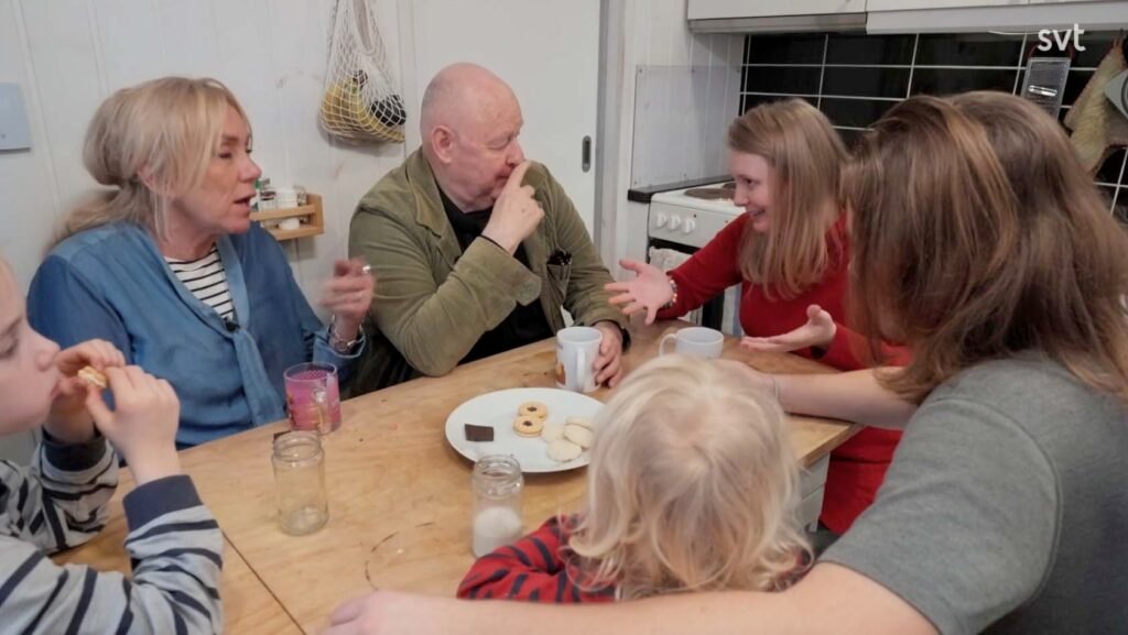 Stiligt - Fem personer, både barn och vuxna, sitter runt ett träbord med koppar och tallrikar med kakor och för en livlig konversation i ett kök.