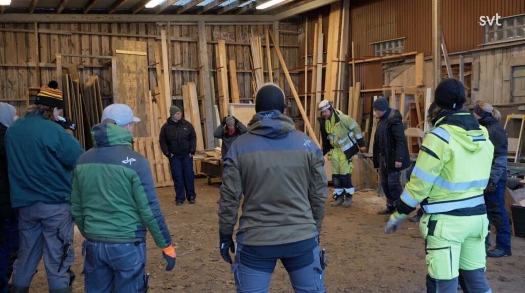 Stiligt - En grupp människor klädda i arbetskläder för utomhusbruk står i en cirkel inne i en verkstad eller ett skjul av trä.