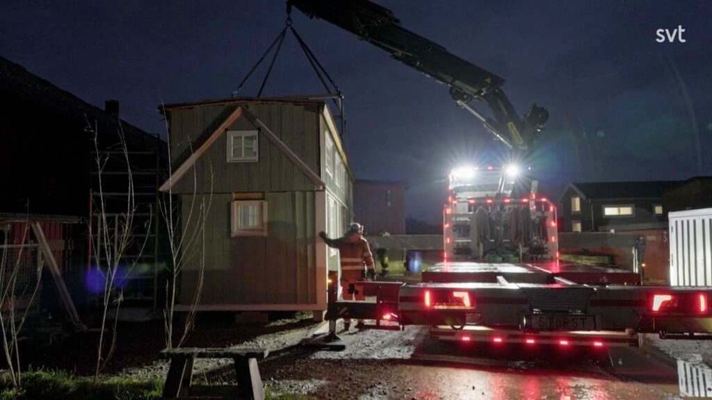 Stiligt - En kran monterad på en lastbil lyfter ett litet växthus medan en person står i närheten och guidar processen i skymningen.