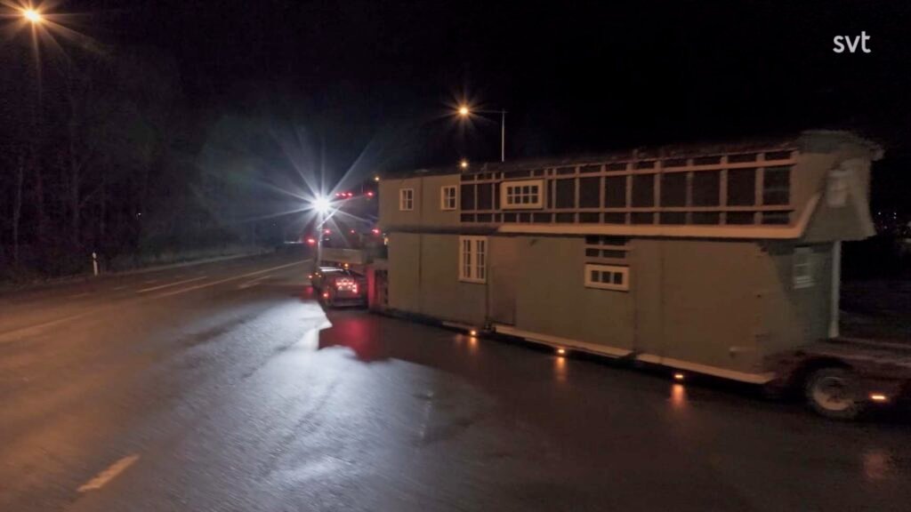 Stiligt - En lastbil transporterar ett stort tvåvåningshus på en släpvagn längs en våt väg på natten, med starka strålkastare i bakgrunden.