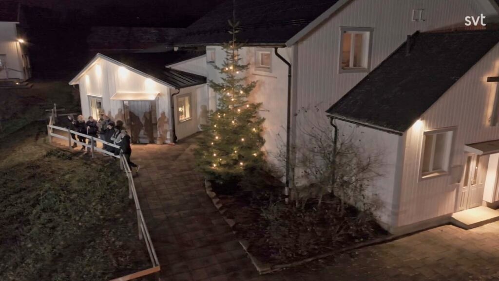 Stiligt - En grupp människor står utanför ett vitt hus dekorerat med en upplyst julgran på kvällen.