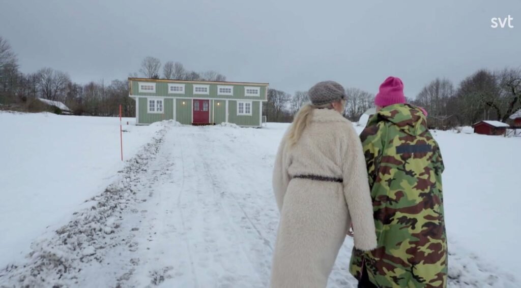 Stiligt - Två personer i vinterrockar går på en snöig stig mot ett grönt hus med röda dörrar, omgivet av kala träd.