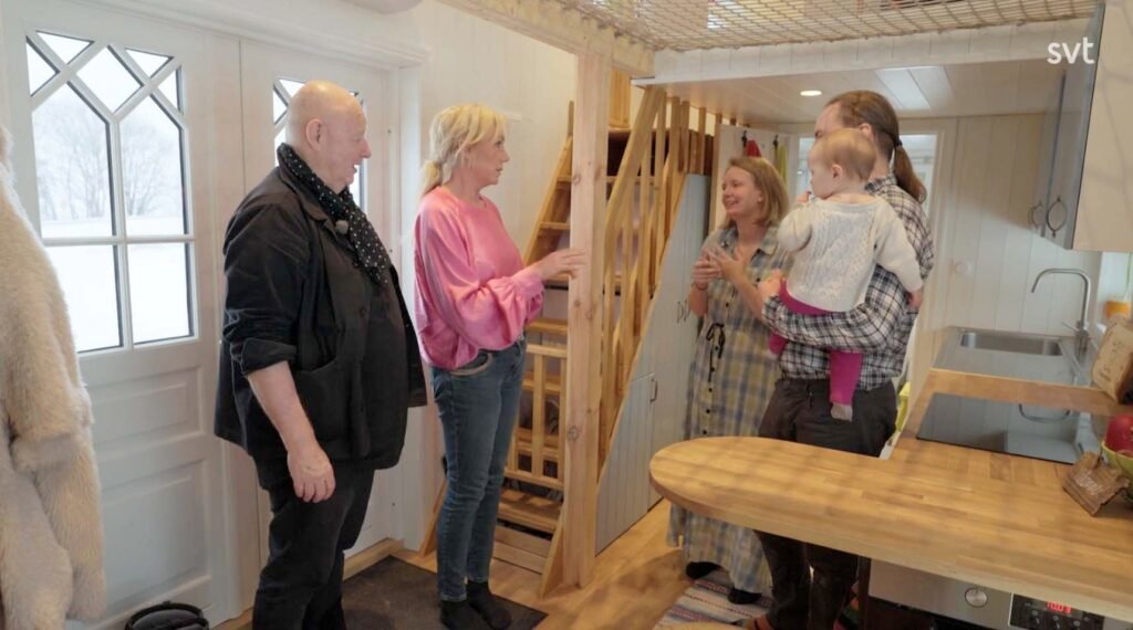 Stiligt - Fem vuxna och en bebis står och pratar i ett litet, modernt trähus med loft och synligt kök.