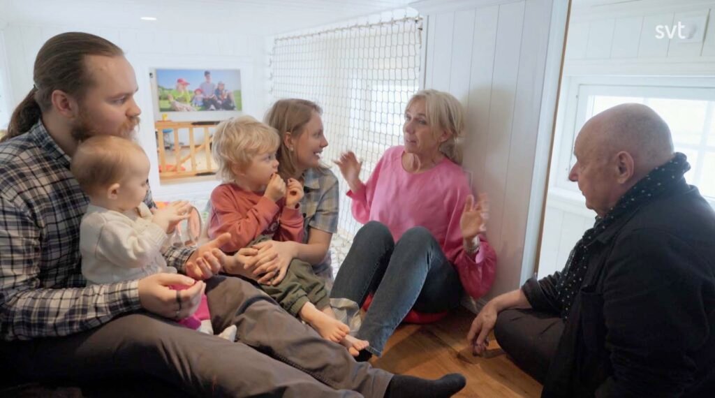 Stiligt - Fem vuxna och två små barn sitter tillsammans i ett litet, ljust rum och samtalar.