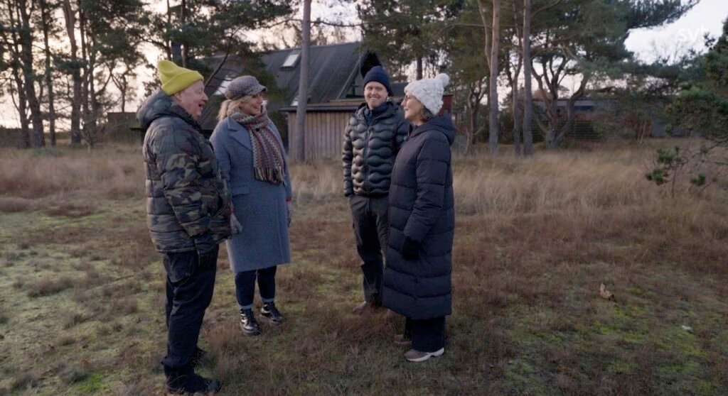 Stiligt - Fyra vinterklädda vuxna står utomhus på en gräsmatta med träd och ett hus i bakgrunden och samtalar.