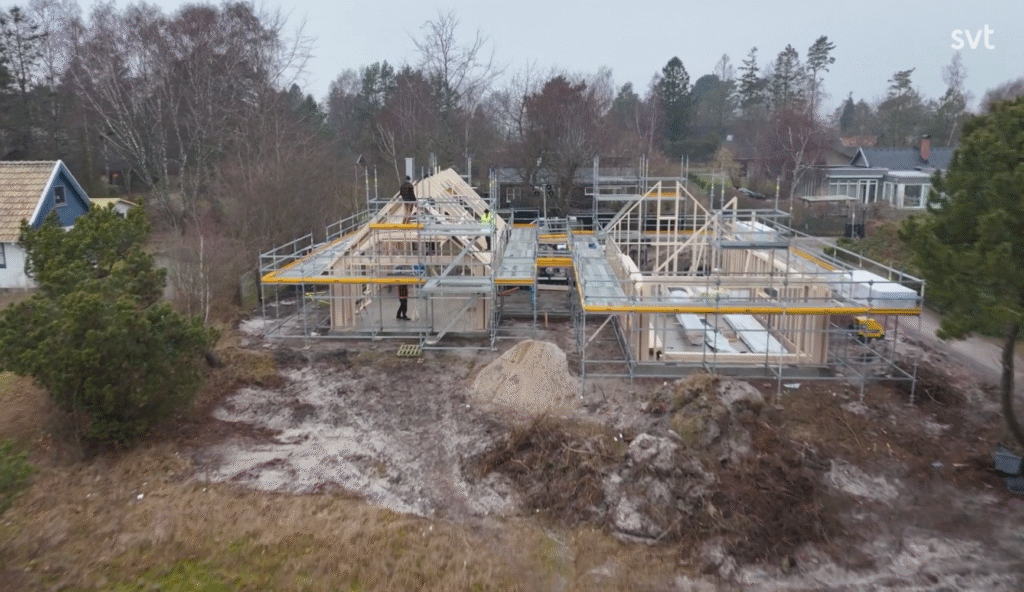 Stiligt - Två hus under uppförande med träramar och byggnadsställningar, omgivna av träd och byggmaterial på en lerig tomt.