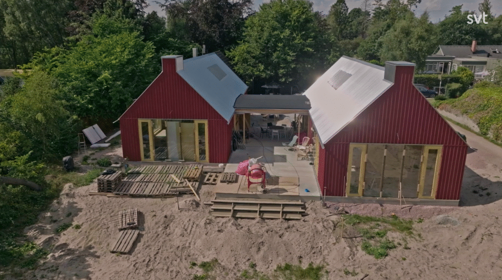 Stiligt - Två moderna röda hus med stora fönster och sluttande plåttak är förbundna med varandra genom en täckt gångväg, omgiven av sand, träpallar och gröna träd.