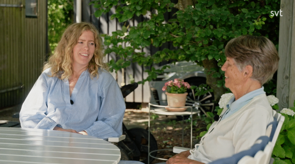 Stiligt - Två personer sitter vid ett runt bord utomhus och samtalar i en lummig trädgårdsmiljö med en krukväxt och skuggande solljus.