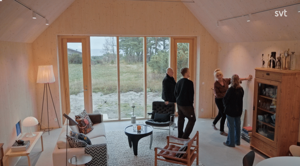 Stiligt - Fem personer står och samtalar i ett modernt, ljust vardagsrum med stora fönster med utsikt över ett gräsbevuxet landskap.