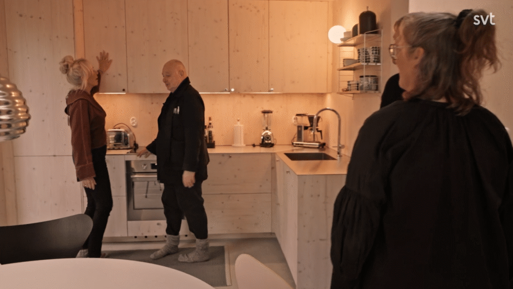 Stiligt - Tre personer står och pratar i ett modernt, ljust träkök; en person står framför spisen, medan de andra står i närheten.