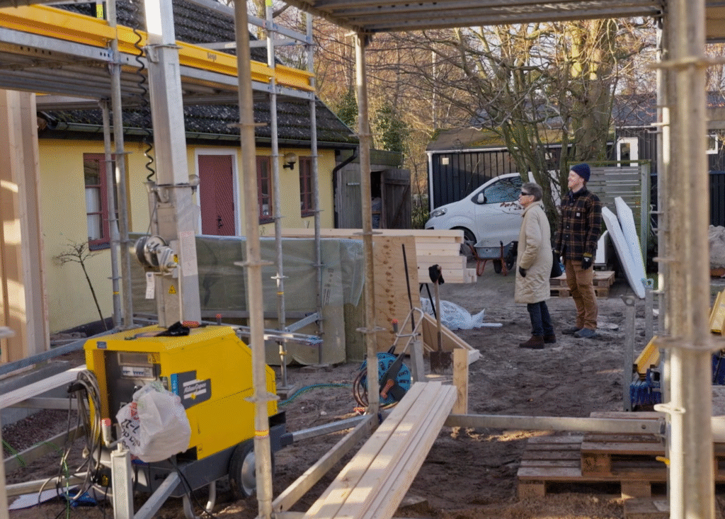 Stiligt - Två personer står bredvid ett hus som håller på att renoveras, omgivna av byggutrustning, träplankor och byggnadsställningar.