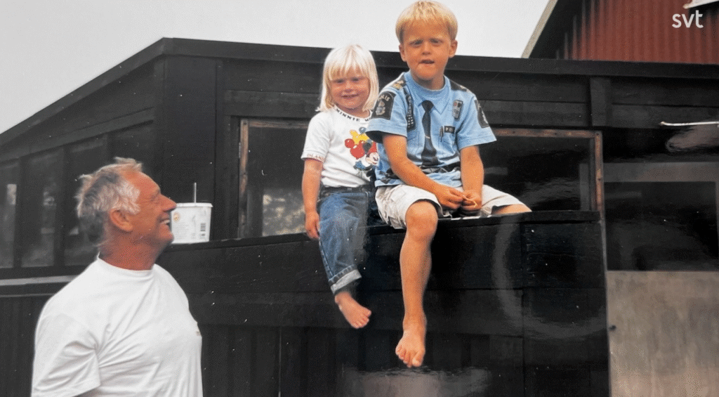 Stiligt - En äldre man står bredvid två barn som sitter på en avsats utanför en mörk träbyggnad, varav ett barn är barfota.