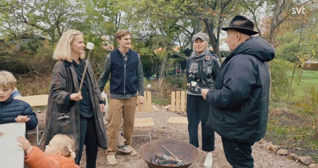 Stiligt - Fem vuxna och två barn står runt en eldstad utomhus, med marshmallows på pinnar, en molnig dag med träd och stolar i bakgrunden.