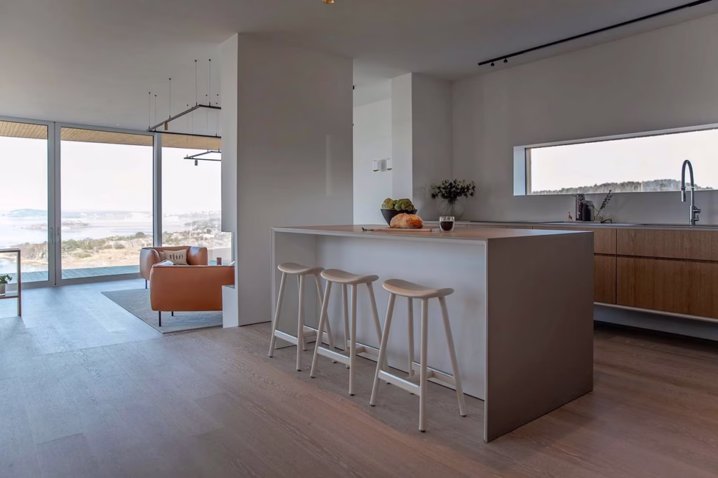 Stiligt - Modernt kök med en stor köksö, tre vita stolar, träskåp och ett vardagsrum med öppen planlösning och stora fönster med utsikt över en naturskön utomhusutsikt.