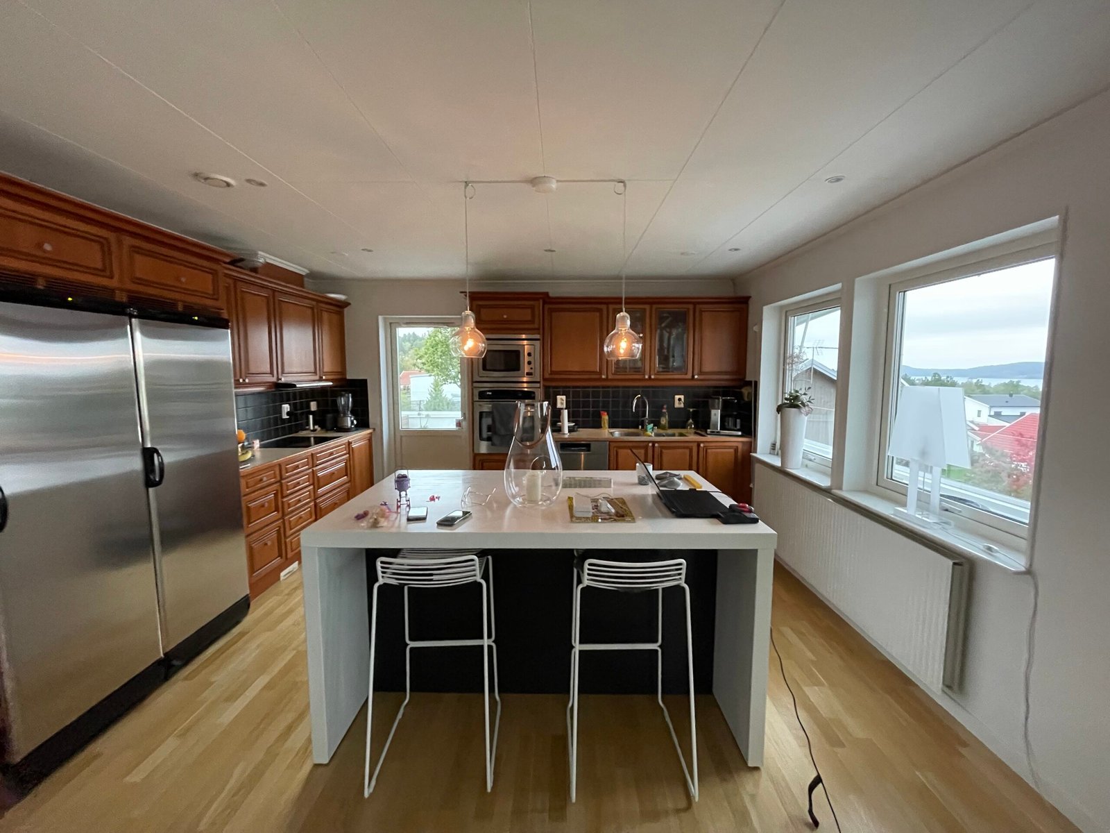 Stiligt - Modernt kök med skåp i trä, rostfria vitvaror, köksö med två vita stolar, pendellampor och stora fönster med utomhusutsikt.