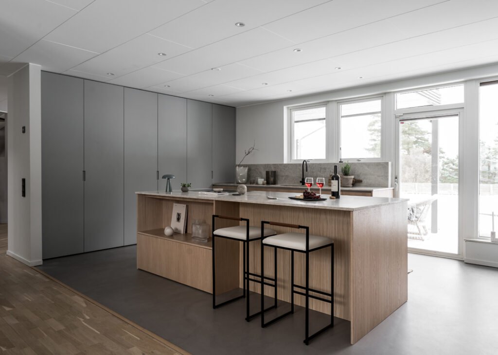 Stiligt - Modernt kök med köksö i ljust trä, två barstolar, grå skåp, inbyggd diskho och stora fönster som släpper in naturligt ljus.