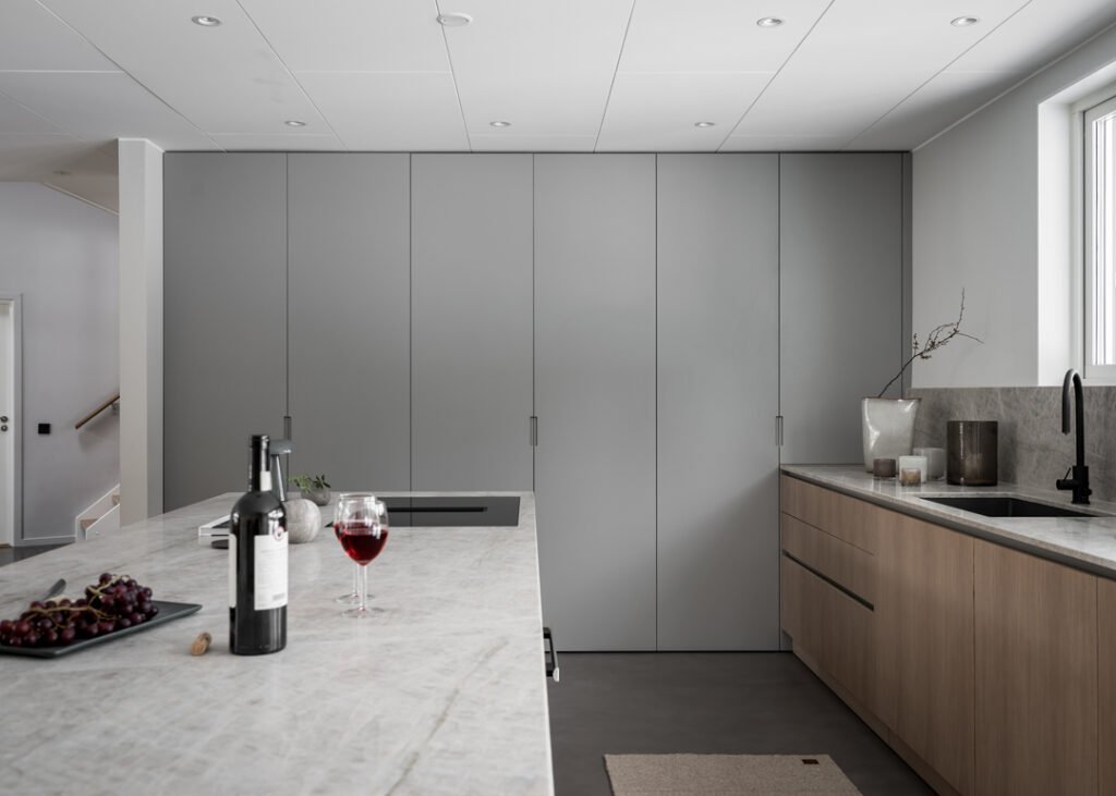 Stiligt - Modernt minimalistiskt kök med grå skåp, bänkskivor i ljus marmor, underskåp i trä, diskho samt en vinflaska och ett glas på köksön. Naturligt ljus kommer in från ett fönster.