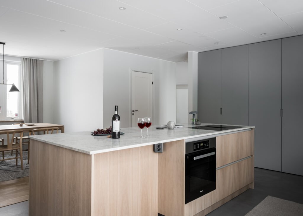 Stiligt - Modernt kök med bänkskiva i marmor, två glas rödvin, en vinflaska, vindruvor, inbyggd ugn och minimalistiska grå skåp; matplatsen syns i bakgrunden.