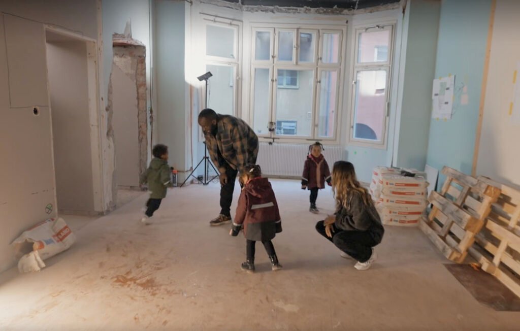 Stiligt - En familj med tre barn står och leker i ett delvis renoverat rum med bara golv, synliga väggar och byggmaterial.