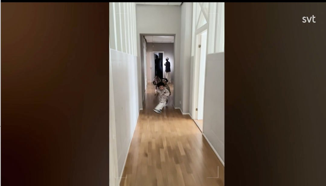 Stiligt - En person i mönstrade kläder sitter på huk i en hall med trägolv, medan en annan person står i bakgrunden. Bilden har "svt" i övre högra hörnet.