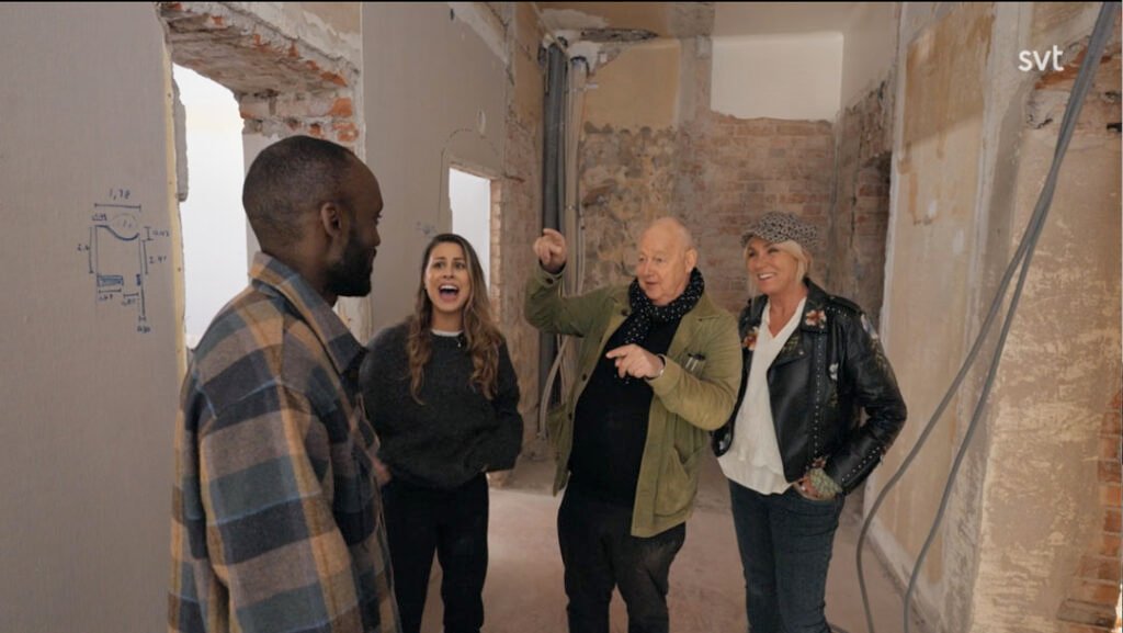 Stiligt - Fyra personer står i ett delvis renoverat rum och pratar och ler bland synliga tegelväggar och byggmaterial.