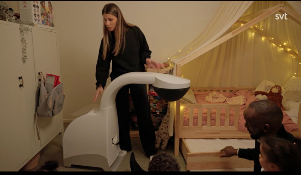 Stiligt - En kvinna manövrerar en stor vit robot i ett barnrum medan en man och två barn sitter på golvet nära en träsäng med ljusslingor.