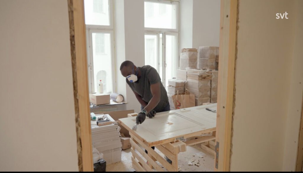 Stiligt - En person iförd mask och handskar arbetar med en dörr i ett rum fyllt av byggmaterial och staplade lådor.