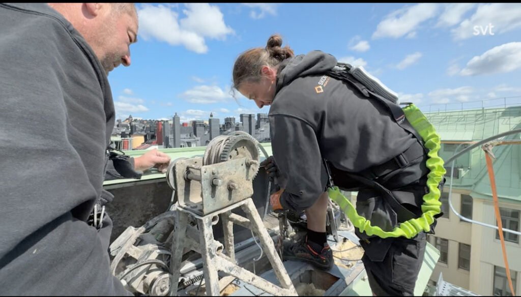 Stiligt - Två arbetare med säkerhetssele manövrerar en vinschmekanism på ett hustak, med stadsbyggnader och en delvis molnig himmel i bakgrunden.