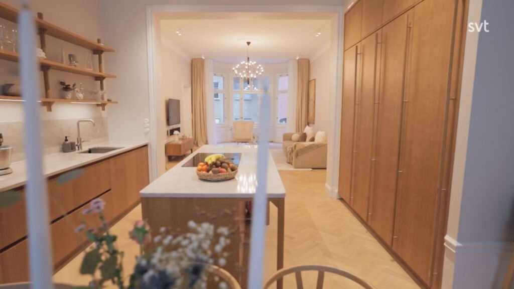 Stiligt - Ett modernt kök med träskåp, en marmorö med en fruktskål, öppna hyllor och utsikt över ett ljust vardagsrum med stora fönster och en ljuskrona.