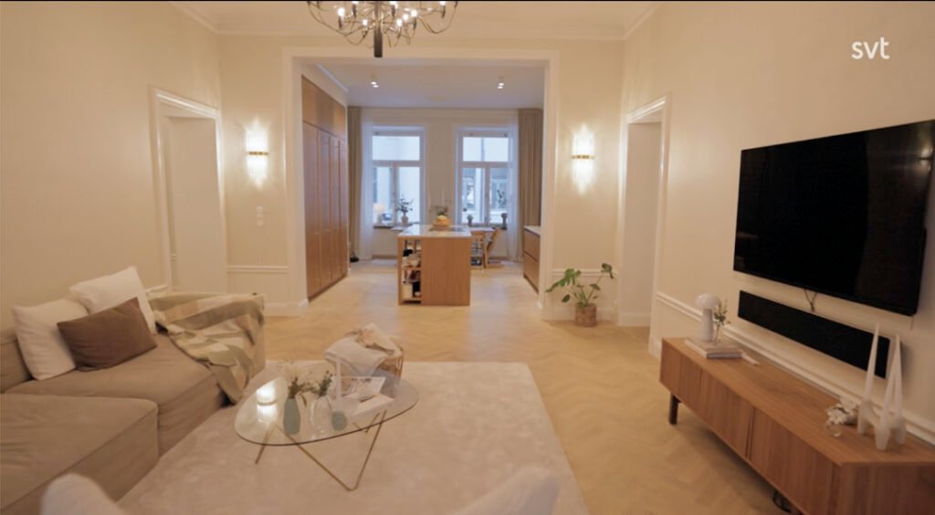 Stiligt - Ett modernt vardagsrum med en beige soffa, trägolv, en väggmonterad TV och en öppen utsikt över en matplats med stora fönster och ett träbord.