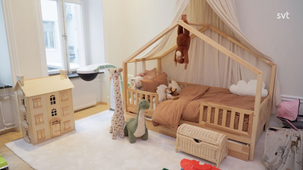 Stiligt - Ett barnrum med en husformad säng av trä, uppstoppade djur, ett leksaksdockhus, en korgkista och en vit matta.