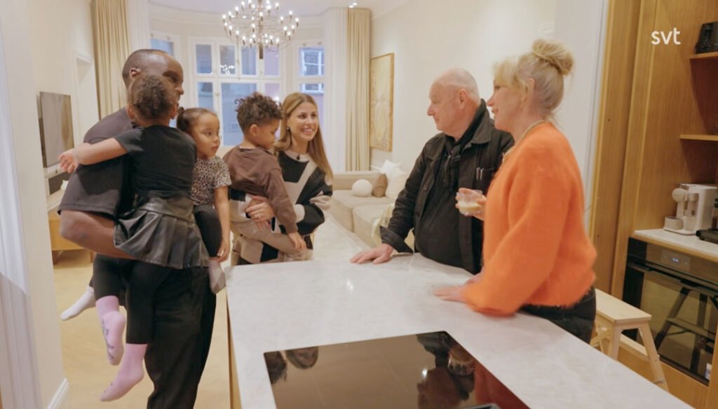 Stiligt - Tre vuxna och två barn står och pratar i ett modernt kök, där en vuxen håller i ett barn och en annan i en drink.