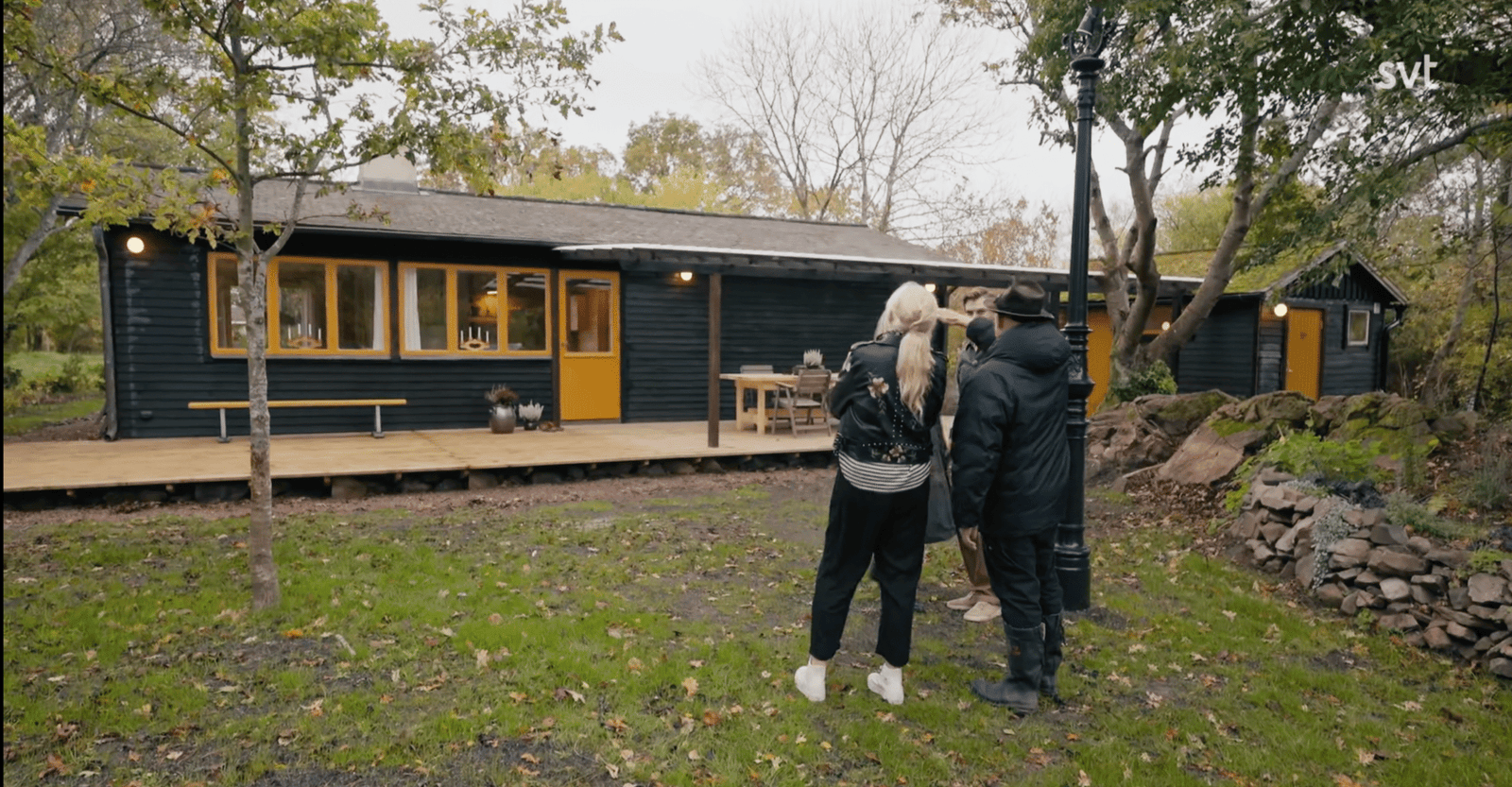 Stiligt - Fyra personer står framför ett mörkt trähus med gula fönsterkarmar och veranda, omgivet av träd och en gräsbevuxen gårdsplan.