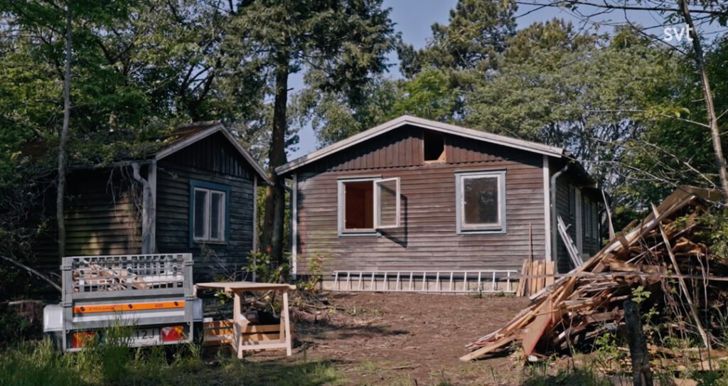 Stiligt - Två små trästugor står bland träden, med en släpvagn, en stege och en hög med träplankor och grenar på gården i klart dagsljus.