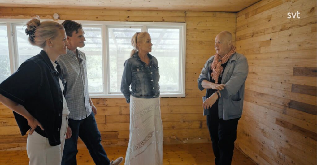 Stiligt - Fyra personer står och samtalar i ett trärum med fönster; en person gestikulerar medan de andra lyssnar.